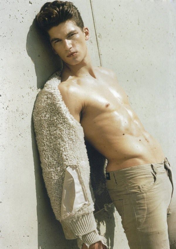 Matteo Guarise leaning on the wall while showing his abs, with a serious face, and wearing a cream fur jacket and beige pants