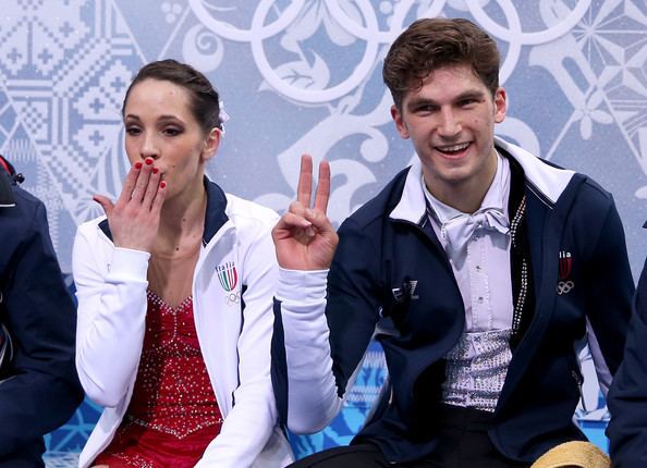 Matteo Guarise smiling with Nicole Della Monica while sitting on the bench. Nicole is giving a flying kiss while Matteo showing a peace sign. Matteo is wearing a white long sleeve with a white bowtie under a white and blue jacket. Nicole is wearing a red dress under a white jacket