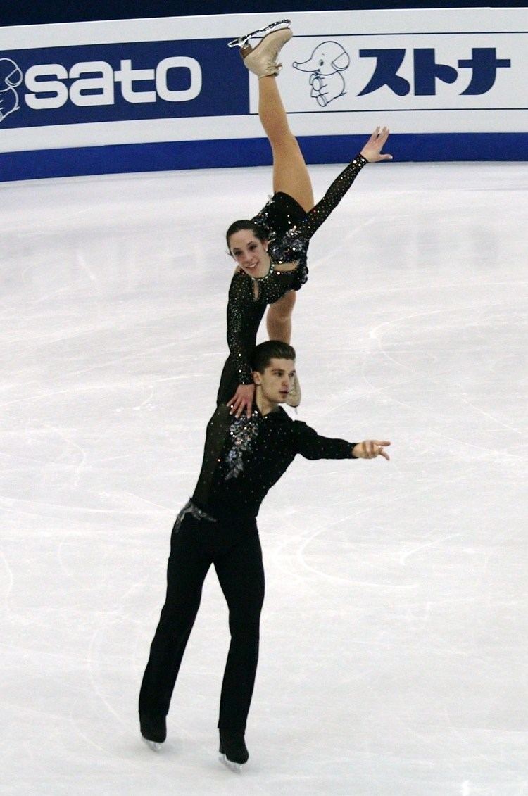 Nicole Della Monica smiling and carried by Matteo Guarise while dancing on the ice. Nicole is wearing a black shiny long sleeve dress  and beige skating shoes while Matteo is wearing black shiny long sleeve, black pants, and black skating shoes
