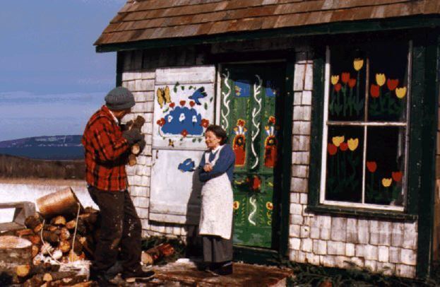 Maud Lewis smiling and talking to man while standing in front of her colorful house. Maud is wearing gray pants, black shoes, and blue long sleeve blouse under a white apron while the man in front of her is wearing a gray beanie, a black and red checkered long sleeve, gray gloves, brown pants, and black shoes