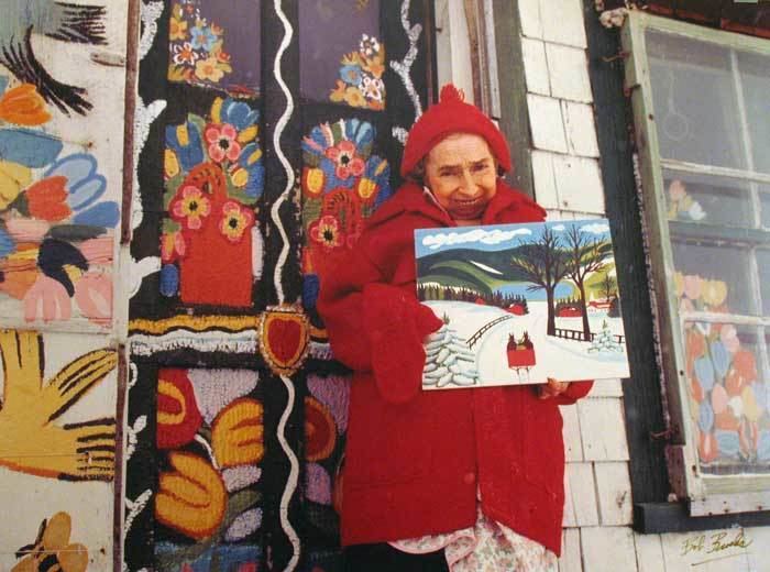 Maud Lewis smiling while holding one of her paintings in front of her Nova Scotia home. Maud is wearing a red beanie and floral dress under a red blazer