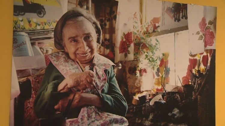 Maud Lewis smiling while sitting on the chair and holding a paintbrush with a painting behind her. Maud is wearing a gray head cover and a green long sleeve blouse under a pink and green floral apron