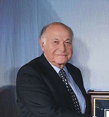 Maurice Tempelsman smiling while wearing a black coat, blue long sleeves, and necktie