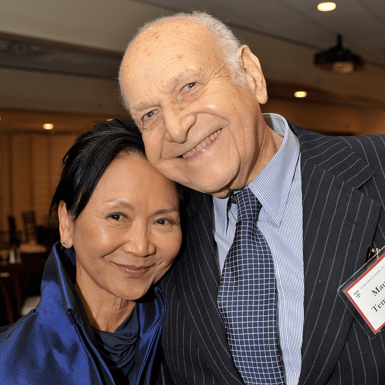 Maurice Tempelsman smiling and wearing a striped coat, long sleeves, and necktie, and the woman beside him is smiling and wearing a blue blouse