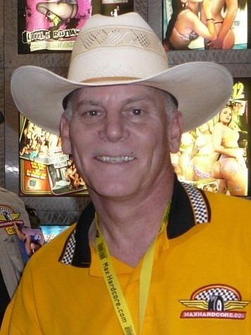 Max Hardcore smiling while wearing a cream cowboy hat, a mustard yellow polo shirt with a black collar, and yellow ID lace