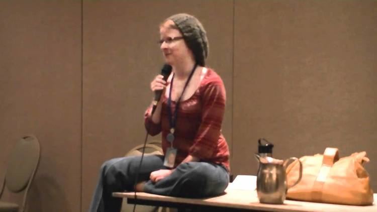 Maxey Whitehead speaking on a microphone while sitting on a table and wearing a red blouse, gray pants