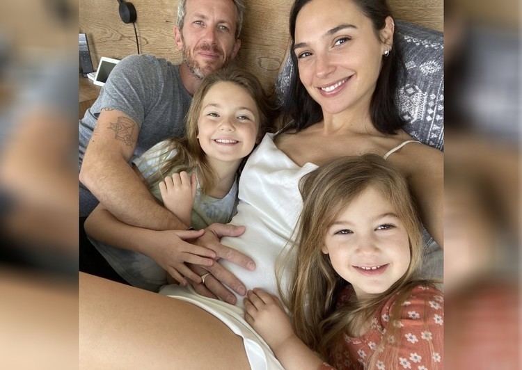 Gal Gadaot smiling in a white dress with her husband Jaron Varsano touching her tummy and their two kids, Maya and Alma