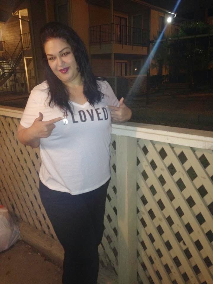 Mayra Rosales smiling in her house and pointing to the "Loved" print on the white shirt she's wearing.