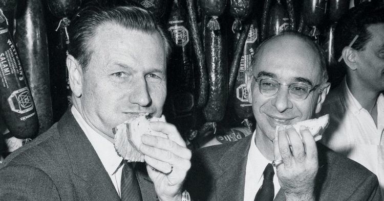 Nelson Rockefeller eating with his friend wearing their suits with a man on their back.