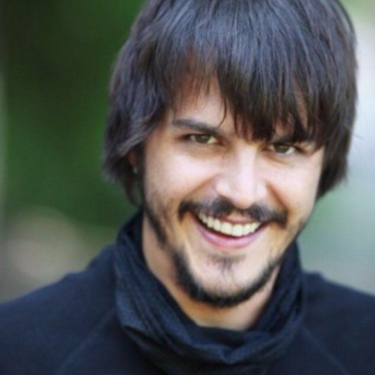 Mehmet Günsür smiling with longer hair and wearing a black scarf and shirt.