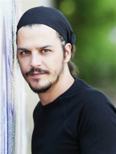 Mehmet Günsür with longer hair leaning into a wall and wearing a black beanie and shirt.