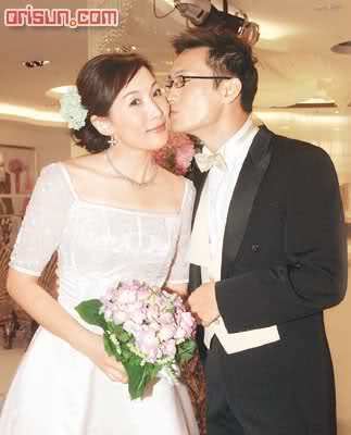 Melissa Ng smiling (left) is smiling while holding a bouquet of flowers, has black hair with a flower clip, wearing silver earrings with a necklace and a white gown. On (right) is a man standing while kissing Melissa on her left cheek,  he is wearing black eyeglasses, black long sleeves, a bow tie under a black suit, and black pants.