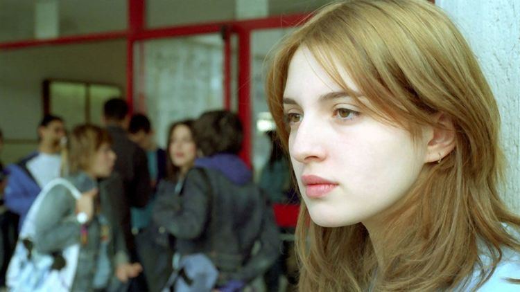 On the left, a group of people talking to each other while on the right, María Valverde looking afar in a scene from the 2005 erotic drama film, Melissa P.