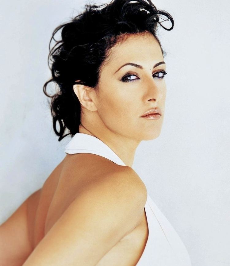 Meltem Cumbul is serious looking to the side, hands on her back is a Turkish actress and TV personality with a mole on the middle of her forehead, and has black curled hair tied up while wearing a white halter, backless dress.