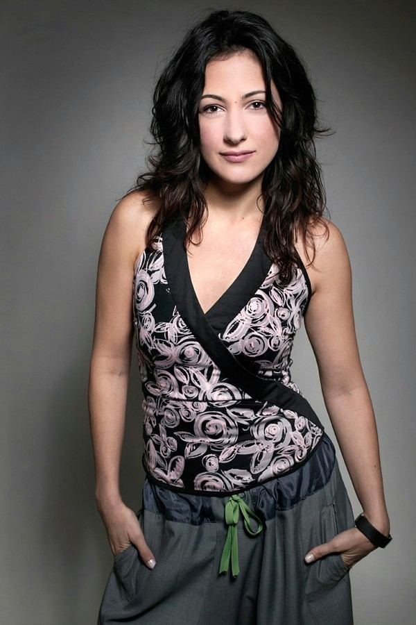 Meltem Cumbul smiling with her hands in her pockets is a Turkish actress and TV personality with a mole on the middle of her forehead and a black curled and messy hair down wearing a black wrist watch on her left wrist while wearing a pair of dark gray harem pants with a green tie, and a black V-neck printed sleeveless top.