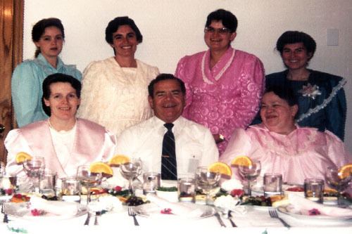Merril Jessop smiling while his six wives are beside him at the dining table and he is wearing white long sleeves and a black necktie