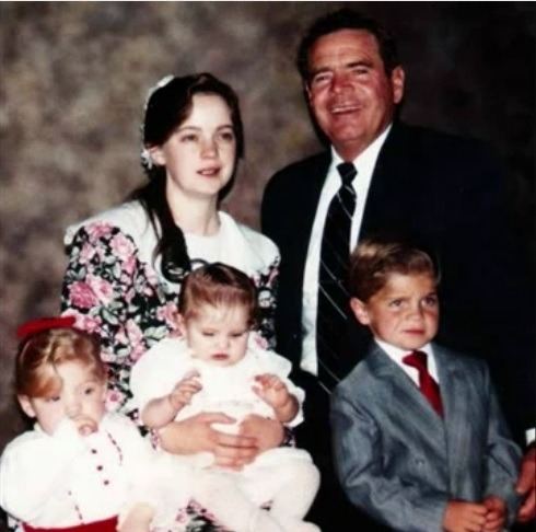 Merril Jessop smiling with his wife and three children while he is wearing a black coat, white long sleeves, and black striped necktie