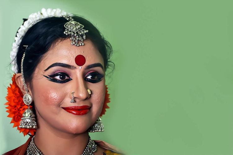 Methil Devika with a tight-lipped smile and make-up on her face while wearing maang tikka, nose-jewel, earrings, and necklace