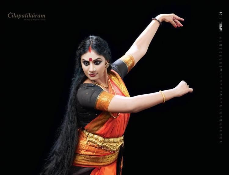 Methil Devika dancing while her eyes are wide open and she is wearing a black and orange dress, gold belt, earrings, bracelet, and layered necklace