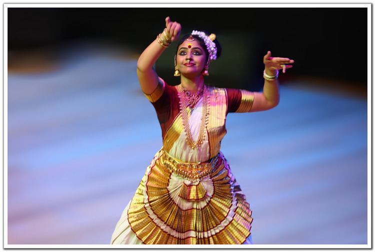 Methil Devika smiling while dancing and wearing a maang tikka, earrings, necklace, bracelet, a maroon and gold blouse, and a white and gold skirt