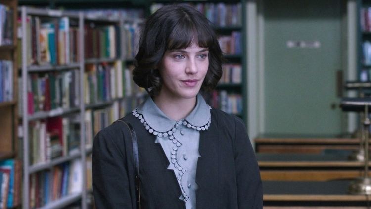 Jessica Brown Findlay smiling while at the library and wearing a black blazer and gray dress in a scene from the 2016 film, This Beautiful Fantastic