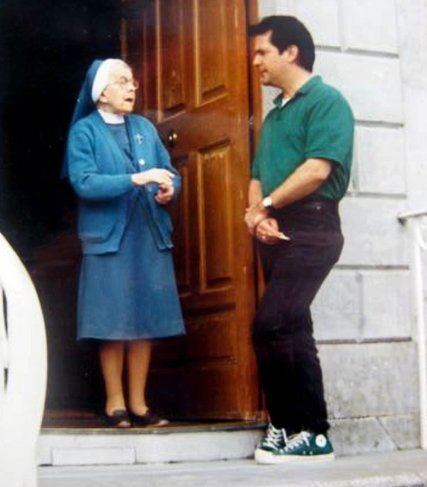 Michael A. Hess wearing a green polo shirt, black jeans, and green shoes with a nun wearing eyeglasses in front of a door.