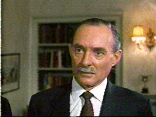 Michael Bates looking serious with a mustache while wearing a white shirt under a black coat and a brown necktie