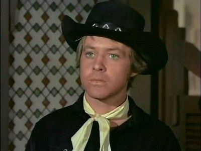 Michael Burns looking serious in a black shirt and black hat with a yellow ribbon on the neck