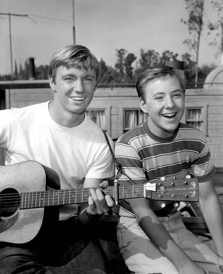 Randy Boone playing the guitar and smiling in a white shirt with Michael Burns wearing a stripe shirt
