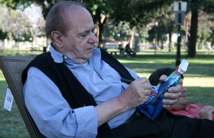 Michael Constantine with a serious face while sitting on a brown chair and writing a spray bottle, wearing a black vest over light blue long sleeves, and black pants.