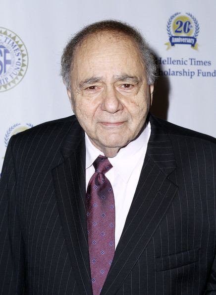 Michael Constantine with a serious face, wearing a black striped coat over white long sleeves, and a purple tie.