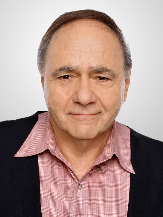 Michael Constantine with a serious face, wearing a black coat over a red checkered polo shirt.