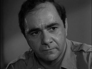 Michael Constantine as Sheriff Charlie Koch with a serious face and wearing a gray polo shirt in a scene from "I am the Night - Color Me Black", a 1964 American television anthology series.