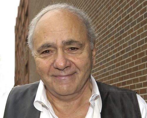 Michael Constantine with a tight-lipped smile, with white hair, wearing a black vest over white long sleeves.