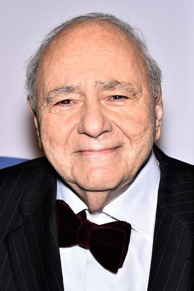 Michael Constantine with a tight-lipped smile, with white hair, wearing a black striped coat over white long sleeves, and a maroon bow tie.