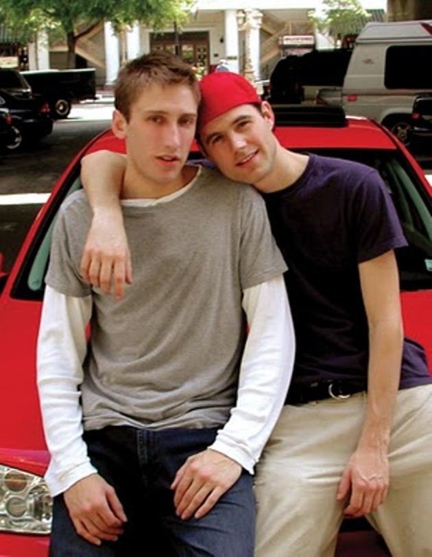 Michael Glatze (left) smiling together with Benjie Nycum (right) while sitting on the hood of a red car with a few other cars in the background. Michael’s hands are resting on his lap, he has brown hair, wearing a white long sleeve under a gray shirt and denim pants while Benjie’s left arm is resting at Michael’s shoulder, he has black hair wearing a red hat, violet shirt and brown pants with a black belt