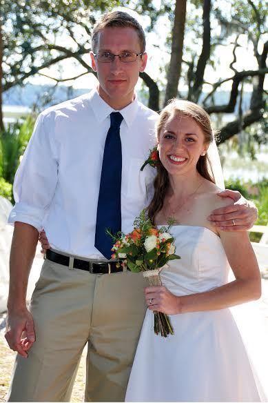 Michael Glatze smiling while touching his left hand on his wife’s shoulder, Rebekah Glatze, with a tree in the background. Michael has blonde hair wearing a ring on his left finger, eyeglass, white polo long sleeve with orange rose on its pocket, a dark blue necktie, and brown pants with a black belt. Rebekah holding a wedding bouquet, she has blonde curly hair, with a white veil wearing a pearl earring, a ring on her left finger, a silver necklace, and a white tube wedding dress