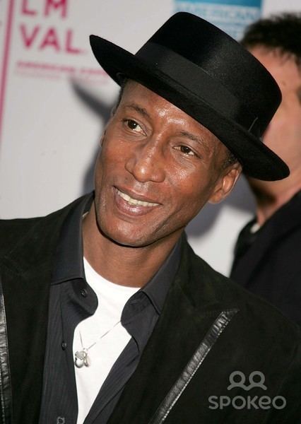 Michael Wright (actor) smiles while wearing a white shirt, black coat and a black hat