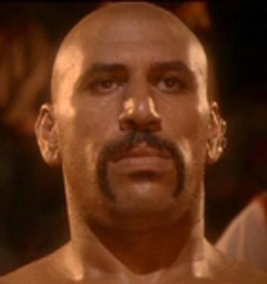 Abdel Qissi as Khan with a serious face, bald hair, a mustache in a movie scene from The Quest, a 1996 American martial arts film.