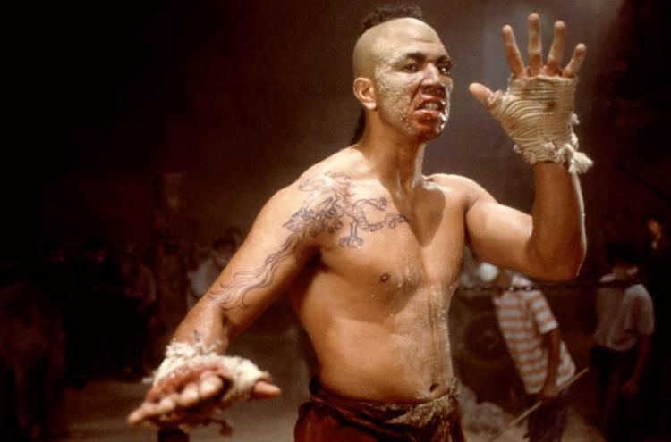Michel Qissi as Tong Po with a furious face, topless, with cloth wrapped on his hands, tattoos on his body, and wearing brown pants in a movie scene from Kickboxer, a 1989 American martial arts film.