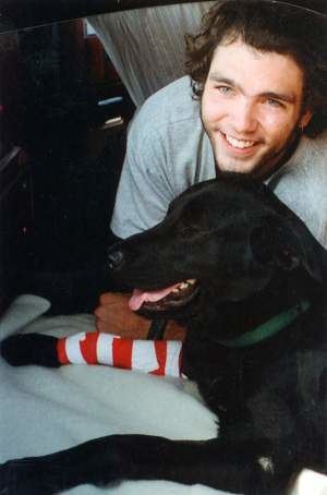 Michel Trudeau smiling with curly hair, beside him is a black dog with a bandage on his foot and his tongue is out while Michel is wearing a gray t-shirt