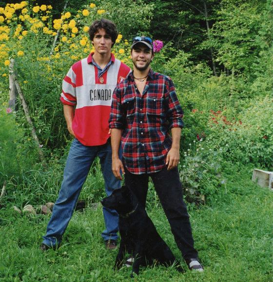 Justin and Michel Trudeau are smiling, behind them are yellow flowers and trees, Justin is wearing a white and red polo shirt and denim pants while Michel is wearing a black and red checkered polo, black pants, and cap