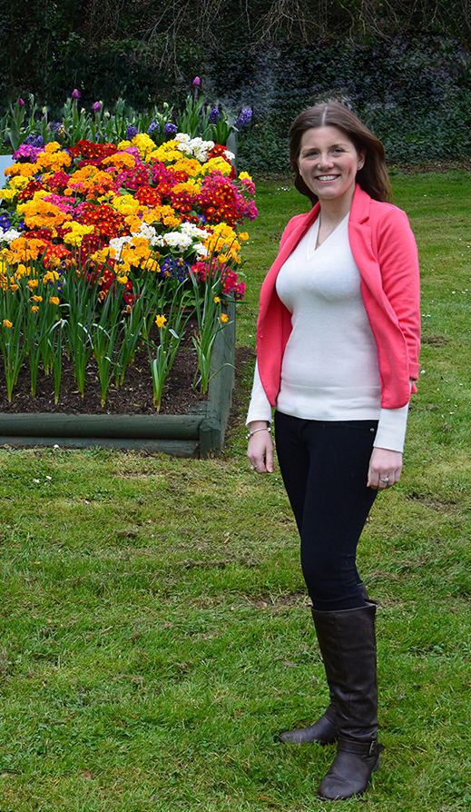 Michelle Donelan smiling while posing at the garden and wearing a pink coat, white long sleeve blouse, black pants, and black boots