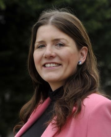 Michelle Donelan smiling while wearing a pink coat, black blouse, and earrings
