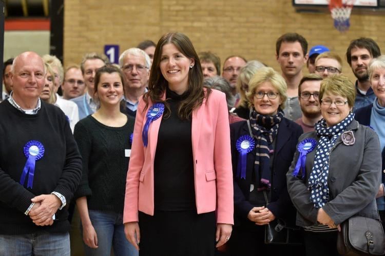 Michelle Donelan smiling with the group of people behind her while she is wearing a pink coat, black dress, and a blue ribbon