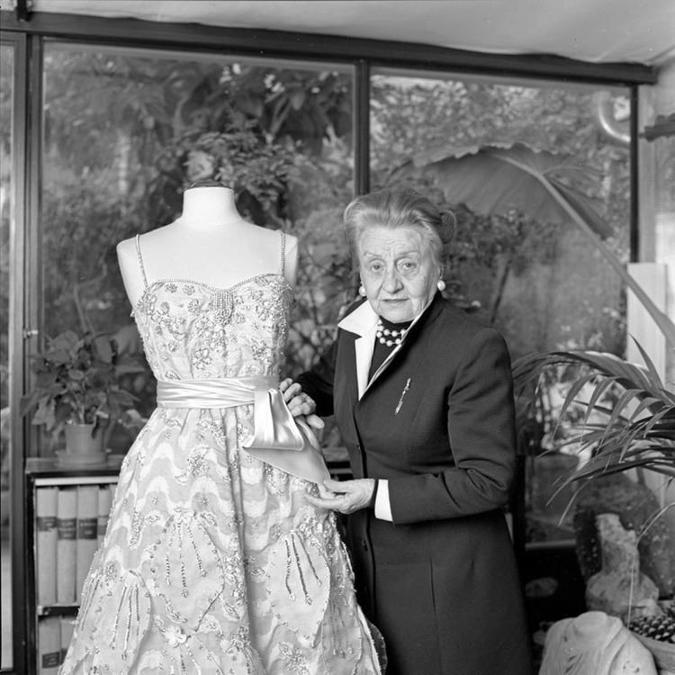 Micol Fontana posing with a gown and wearing a collared dress.
