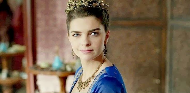 Pelin Karahan as Mihrimah Sultan with a tight-lipped smile while wearing a blue gown, a crown, and necklace in the 2011 tv series, The Magnificent Century