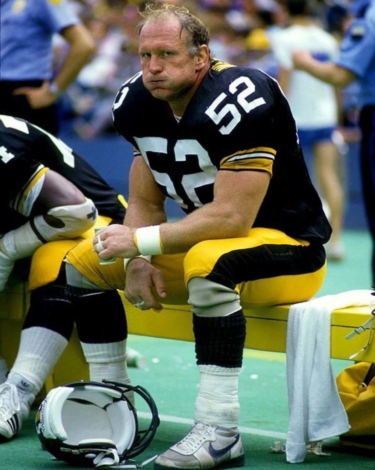Mike Webster's mouth is full of water while sitting on the bench, holding a paper cup, and wearing a black, white, and yellow football jersey with number fifty-two