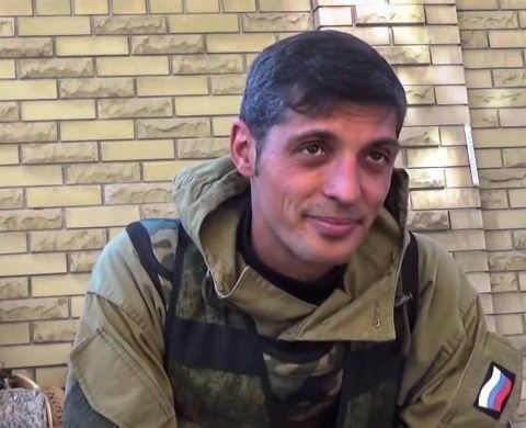Mikhail Tolstykh with a tight-lipped smile and looking at something while wearing a brown jacket and camouflage tactical vest