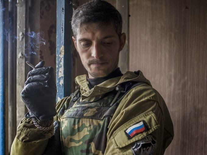 Mikhail Tolstykh smoking and looking down, with a mustache, while wearing a brown jacket, camouflage tactical vest, black gloves, and bracelet